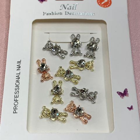 A package of nail art decorations includes silver, gold, and rose gold metallic teddy bear shapes, each adorned with a heart-shaped rhinestone. The packaging is labeled "Teddy Bear Charms with Crystal Rhinestone Heart for Nails Art Design" by Dynamic Nail Supply and features butterfly icons in the lower right corner.