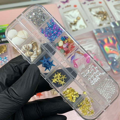 A hand clad in a black glove showcases the Dynamic Nail Supply's "Summer Nail Charm," a clear plastic case filled with vibrant nail art decorations, including seashells, starfish, beads, faux gems, and pearls. In the background, an array of colorful nail supplies is spread across a soft pink surface.