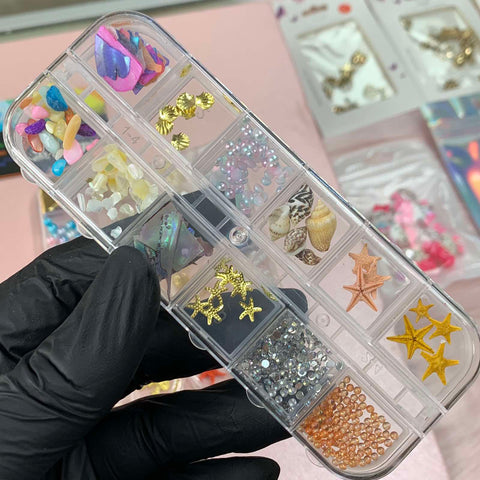 A hand in a black glove holds a transparent organizer filled with vibrant summer nail art decorations from Dynamic Nail Supply. The compartments are adorned with colorful seashell and starfish charms, along with beads and gems. In the background, more essential nail supplies await creative inspiration.