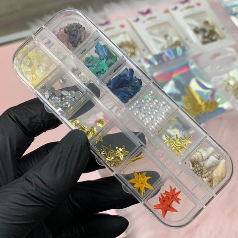 A gloved hand holds a clear plastic organizer filled with Dynamic Nail Supply's "Summer Nail Charm," which includes vibrant rhinestones, seashells, starfish sequins, and pearly accents—ideal for creating summer nail art designs. In the background, more nail supply packages are displayed on a pink surface.