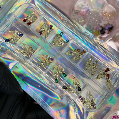 A person wearing black gloves holds a holographic package from Dynamic Nail Supply containing 24 spike-shaped nail charms, embellished with jewels. These designs resemble lightning bolts and are adorned with sparkling stones in vibrant colors such as red, blue, green, and black. Perfect for any nail charm enthusiast!