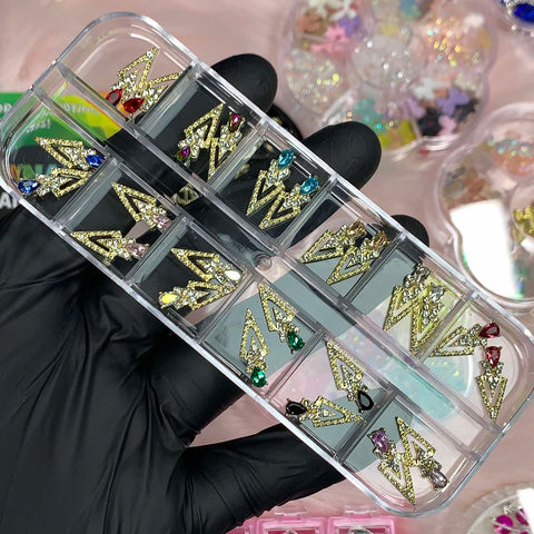 A gloved hand holds a clear rectangular box showcasing an assortment of sparkly, spike-shaped earrings, each adorned with colorful gemstones in their own compartments. In the background, tools and nail charms from Dynamic Nail Supply are scattered among other nail art supplies and decorations.