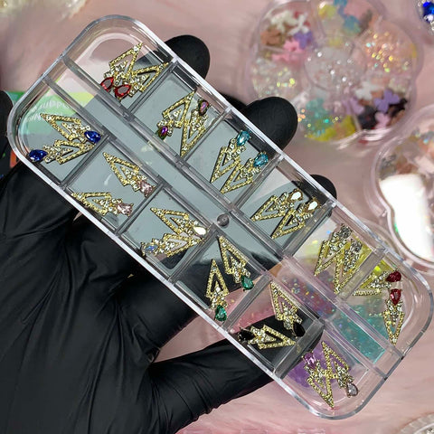 A gloved hand presents a clear case with gold, spike-shaped nail charms from Dynamic Nail Supply's collection. The case is filled with various charms adorned with vibrant gemstones in shades of blue, red, and green. In the background, blurred jars containing small, colorful decorations can be seen.