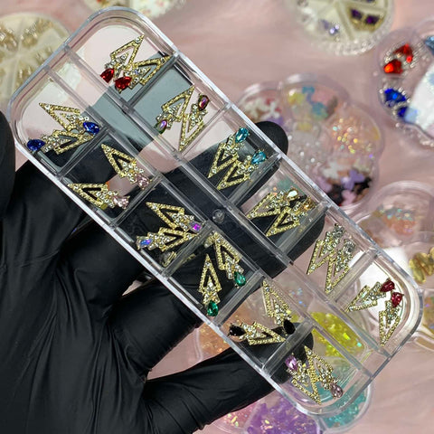 In a gloved hand with exquisite nail art, a transparent case showcases the Dynamic Nail Supply's collection of 24 spike-shaped charms for designers, each adorned with colorful gemstones. The charms feature gold accents and are neatly arranged in compartments, while blurred trays filled with more jewelry pieces create a soft pink backdrop.