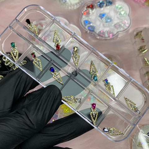 A gloved hand holds a clear jewelry organizer showcasing six pairs of gold-toned triangular earrings, each embellished with colorful gems. Accentuating the vibrant red, blue, green, and clear stones are spike-shaped nail charms from Dynamic Nail Supply's 12 pcs collection for designers. In the background, trays filled with more colorful gems are set against a pink surface.