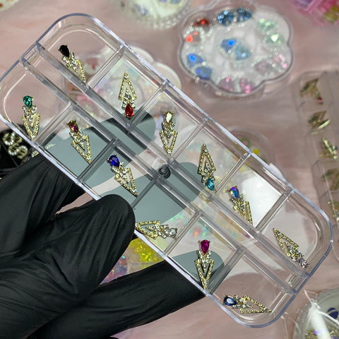 A gloved hand holds a transparent container featuring spike-shaped nail charms from Dynamic Nail Supply, showcasing their array of twelve gold spikes adorned with various colored gemstones. The soft pink background is decorated with trays filled with colorful beads and decorative gems, ready to inspire a nail art designer.
