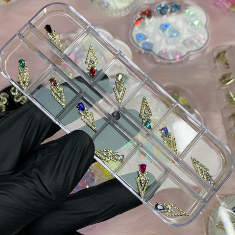 A gloved hand holds a clear container showcasing various earrings, each featuring a triangular gold design with a colored gemstone at the center. The earring designs are accentuated by subtle embellishments from the 12 pcs spike-shaped Nail Charms by Dynamic Nail Supply, while additional decorative pieces are visible on a pink surface in the background.