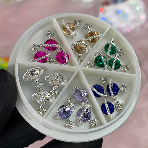 The Dynamic Nail Supply 18 pcs Planet Charms for Nails designer features a round white container divided into six sections, each containing a pair of sparkly earrings adorned with silver accents and colorful gemstones in pink, amber, green, clear, purple, and blue. A gloved hand gracefully holds it against a blurred pink background.
