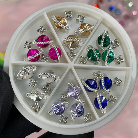 Introducing the 18 pcs Planet Charms for Nails by Dynamic Nail Supply: a stylish round white container containing six compartments. Each slot is filled with vibrant gemstones set in intricate metal settings, perfect for trendy nail art enthusiasts. The gemstones are arranged in pairs showcasing vivid colors such as pink, green, blue, lavender, gold, and clear. Accompanying each pair are delicate flower-shaped metal accents.
