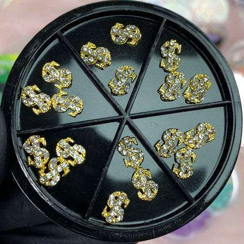 A gloved hand supports an elegant black circular container from Dynamic Nail Supply, designed for nail art enthusiasts. Inside, the container is sectioned into six parts, each filled with multiple pieces of gold-colored Small Dollar Sign Charms adorned with shimmering rhinestones. This setup effortlessly merges luxury and creativity against a softly colored background.