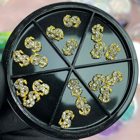 The 18-piece set of small dollar sign charms for nails from Dynamic Nail Supply comes in a black circular container with nine sections, offering both gold and silver options ideal for nail art enthusiasts. Each charm is adorned with small sparkling stones and is elegantly displayed within the compartments on a gloved hand.