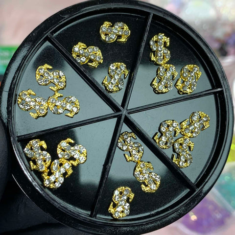 An 18-piece set of small dollar sign charms from Dynamic Nail Supply is housed in a round black container with eight compartments. The gold and silver rhinestone-encrusted charms glisten under the light, featuring a mix of metallic and shiny crystal elements—a must-have for dazzling nail art enthusiasts.