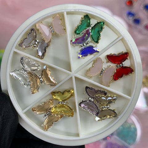 A circular container featuring eight compartments, each filled with one of the vibrant 12 pcs Butterfly Rhinestone Charms for Nails Art Designer, showcasing a range of colors including pink, clear, green, blue, red, beige, yellow, and gold. A black-gloved hand displays the Dynamic Nail Supply container against a blurred backdrop.