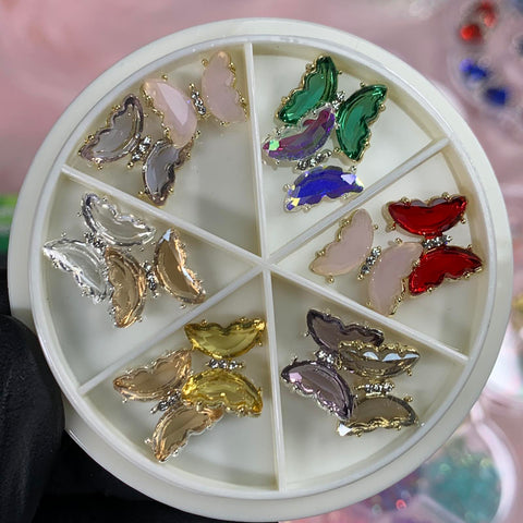 A gloved hand holds a white circular container from Dynamic Nail Supply against a background of colorful blurred objects. This artistic Nails Art Designer piece features six compartments, each filled with vibrant butterfly rhinestone charms in silver, gold, pink, green, red, and purple.