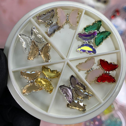 A round container from Dynamic Nail Supply contains the 12 pcs Butterfly Rhinestone Charms for Nails Art Designer in pink, purple, green, yellow, and red. Each butterfly features metallic edges and is adorned with rhinestones. A gloved hand holds the container steady as the background appears blurred.