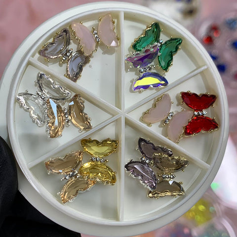 A close-up of a circular white organizer containing six sections, each filled with the vibrant 12 pcs Butterfly Rhinestone Charms for Nails Art Designer by Dynamic Nail Supply. These exquisite charms come in light pink, green, red, gold, silver, and translucent colors. The stunning collection is showcased against the backdrop of a hand wearing black gloves.