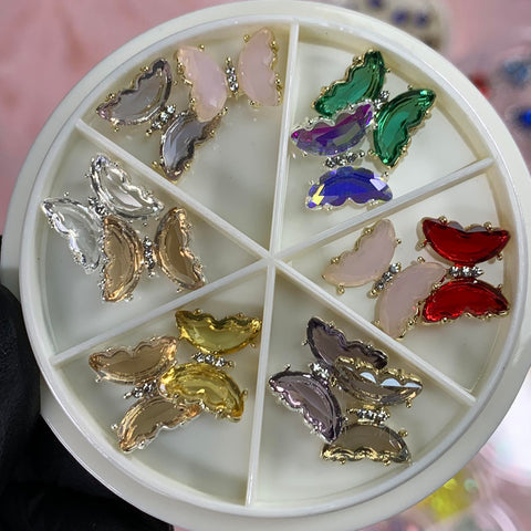 A circular container holds six compartments of decorative nail charms, ideal for any nails art designer. These rhinestone-adorned accessories feature butterfly designs in dynamic colors such as pink, green, red, silver, gold, and purple. Brought to you by Dynamic Nail Supply.
