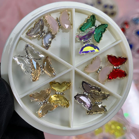 A gloved hand holds the Nails Art Designer's creation from Dynamic Nail Supply: a round white container featuring six sections, each containing butterfly-shaped charms in vivid colors, including pink, green, red, gold, clear, and purple. The background is beautifully blurred.