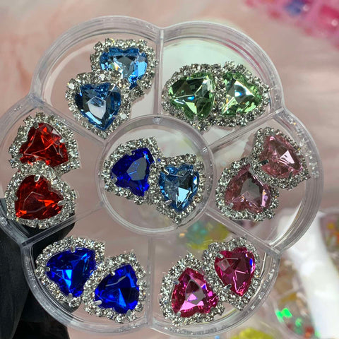 A round, transparent plastic container from Dynamic Nail Supply encases the 14 pcs Heart Shaped Rhinestone Charms for Nails Art Designer. Each charm boasts a central gem in striking colors such as blue, red, green, and pink, bordered by tiny clear rhinestones. The container is sectioned into compartments to showcase these dazzling charms for nail art designers.