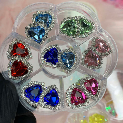A gloved hand elegantly displays a circular container from Dynamic Nail Supply, designed to hold heart-shaped rhinestone charms, perfect for any nails art designer. The container features six sections, each showcasing a different color: red, light blue, green, dark blue, pink, and magenta. Outlined with small silver rhinestones from their 14 pcs Heart Shaped Rhinestone Charms collection.