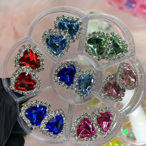 The 14 pcs Heart Shaped Rhinestone Charms for Nails Art Designer from Dynamic Nail Supply is a circular plastic container divided into six sections, featuring vibrant rhinestones ideal for nail art designers. Each section contains heart-shaped rhinestone charms in shades like blue, green, red, and pink, accented by small clear stones on a soft pink background.