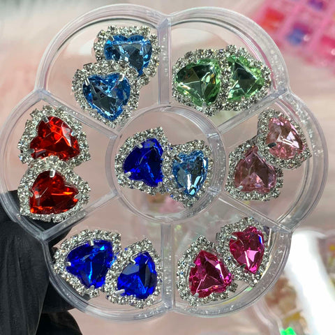 A clear flower-shaped organizer from Dynamic Nail Supply elegantly showcases the 14 pcs Heart Shaped Rhinestone Charms for nails art designers, each adorned with silver rhinestone borders. The gems, available in vibrant shades of red, blue, green, and pink, are neatly arranged in separate compartments. A black-gloved hand presents the organizer against a softly blurred backdrop.