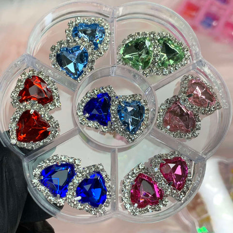 A circular container from Dynamic Nail Supply houses sparkling, heart-shaped gemstones in an array of colors, including red, blue, green, and pink. Each gemstone is encircled by clear rhinestones. Ideal for Nails Art Designers seeking Heart Shaped Rhinestone Charms, the gems are organized neatly within separate sections of the container.