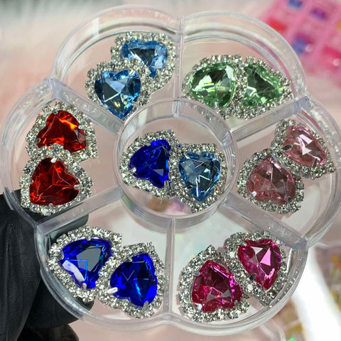 A black-gloved hand holds a round container of Dynamic Nail Supply's 14 pcs Heart Shaped Rhinestone Charms for Nails Art Designer, showcasing an assortment of colorful gems—red, blue, green, and pink—each surrounded by small, sparkling stones. Ideal for nail art designers, this collection features elegant heart-shaped charms.