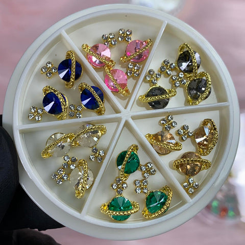 A gloved hand holds the Dynamic Nail Supply container, a round organizer featuring their "18 pcs (Gold) Planet Charms for Nails designer." Each compartment showcases pairs of decorative nail art charms adorned with colorful, teardrop-shaped gems in hues of blue, pink, black, clear, green, and amber. These gems are elegantly set in gold and accompanied by small clusters of clear rhinestones.