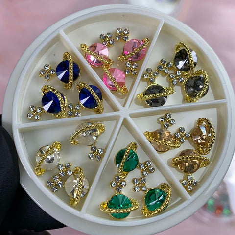 A round container featuring six compartments beautifully displays the Dynamic Nail Supply's 18 pcs (Gold) Planet Charms for Nails designer. Each pair includes a large gemstone in pink, blue, green, or clear, surrounded by golden accents and small rhinestones. A black-gloved hand elegantly supports this stunning showcase of nail art wonders.