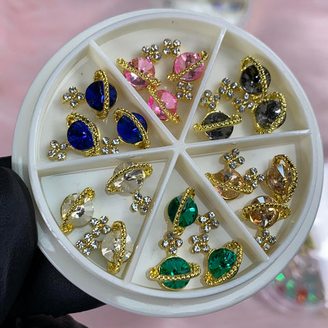 A gloved hand holds a circular plastic container against a light pink background, showcasing Dynamic Nail Supply's 18-piece collection of teardrop-shaped Planet Charms for Nails. Each charm, surrounded by sparkling stones, features vibrant shades of blue, pink, black, clear, and green—a true nail art delight.