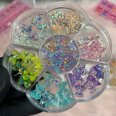 A gloved hand holds a round, transparent plastic organizer containing an assortment of colorful, iridescent butterfly-shaped beads perfect for fashionable nail art designs. These beads come in shades of yellow, green, blue, purple, pink, and clear. The center section is filled with small multicolored beads.