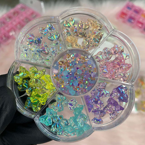 Introducing the 60-piece 3D Resin Bear Charms for Nails, specifically designed for nail art enthusiasts by Dynamic Nail Supply. This clear circular container, ideal for any nail art designer, is divided into seven sections that are filled with trendy tiny charms in colors such as clear, yellow, blue, green, purple, pink, and iridescent. At its center lies a collection of small rhinestones. The background highlights subtly blurred pastel-colored boxes against a soft and fuzzy surface.