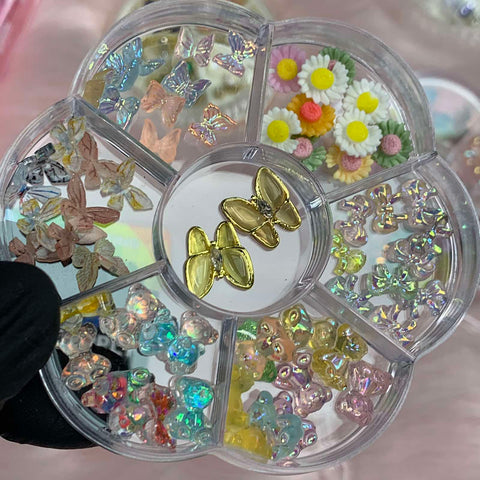A gloved hand holds the circular organizer by Dynamic Nail Supply, featuring a flower-shaped design with multiple compartments against a blurred backdrop. It contains an assortment of decorative nail art pieces from their 60 pcs Resin Butterfly & Teddy Bear & Bow & Flower Charms with Rhinestones for Nails Art Designer collection, including resin butterfly charms, daisies, and various colorful, iridescent beads.
