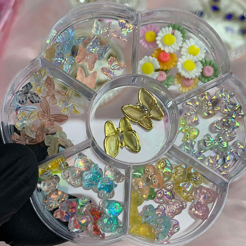 A circular container from Dynamic Nail Supply offers a delightful array of decorative nail art items across six sections, featuring colorful resin butterfly, teddy bear, bow, and flower charms adorned with rhinestones. Translucent butterflies, flowers, and iridescent shapes in pastel hues fill each section. At the center is an eye-catching larger butterfly design held elegantly by a hand wearing a black glove.