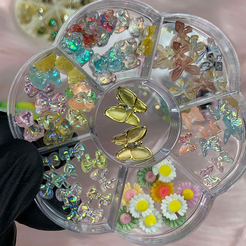 A gloved hand holds a circular compartmentalized container from Dynamic Nail Supply against a soft pink background, showcasing their set of 60 colorful nail art embellishments. The sections feature iridescent resin butterfly charms, pastel flowers, shiny stars adorned with rhinestones, and golden bows.