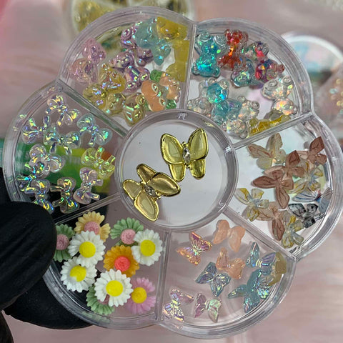 A flower-shaped container from Dynamic Nail Supply is perfect for a nail art designer, featuring sections filled with colorful resin butterfly and teddy bear charms, golden bow-shaped pieces in the center, pastel floral designs with daisies, and rhinestone embellishments. It’s held by a gloved hand against a soft pink background.