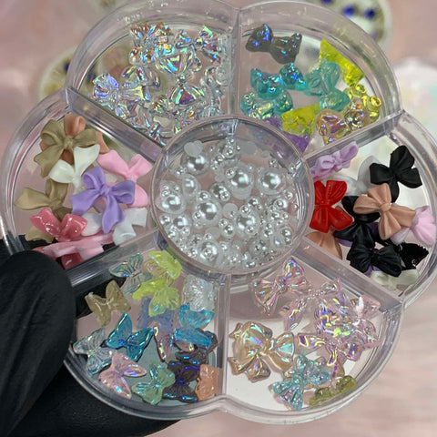 A round plastic organizer from Dynamic Nail Supply, labeled "60 pcs Mix-shapes Resin Charms with Pearls for Nails Art Designer," elegantly showcases a charming collection of nail art essentials. Five compartments are filled with vibrant, bow-shaped resin charms, while the center compartment features white pearl-like beads. The piece is gracefully displayed by a gloved hand against a softly blurred background.