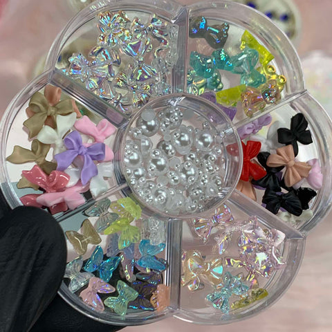 A gloved hand holds the clear, flower-shaped container from Dynamic Nail Supply against a blurred background. Inside, the 60-piece set of mixed-shape resin charms for nail art includes colorful bow-shaped designs and iridescent beads in brown, pink, purple, blue, yellow, and black.
