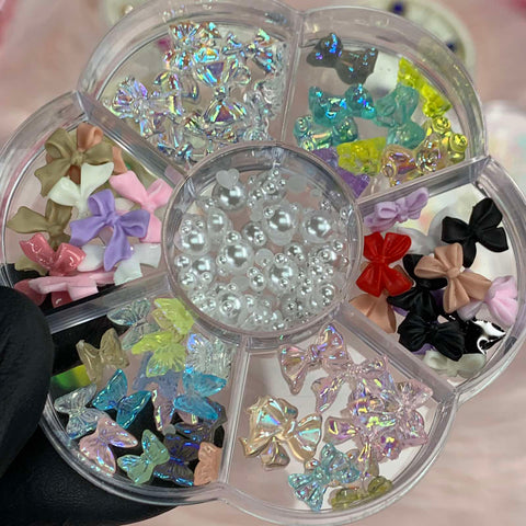 A black-gloved hand elegantly showcases the Dynamic Nail Supply treasure: the "60 pcs Mix-shapes Resin Charms with Pearls for Nails Art Designer." This flower-shaped container holds a variety of decorative items perfect for nail art, featuring pearl-like beads at the center and butterfly-shaped resin charms in colors such as pink, purple, black, green, and clear.