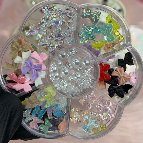 A gloved hand holds a beautifully crafted flower-shaped container from Dynamic Nail Supply, featuring the "60 pcs Mix-shapes Resin Charms with Pearls for Nails Art Designer." This elegant organizer has eight compartments, showcasing an assortment of decorative nail art pieces: small pearls occupy one section, while other sections display resin charms and colorful plastic bows in various shades such as purple, pink, blue, clear, yellow, orange, black, and red.