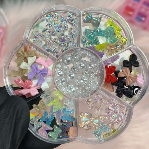 A black-gloved hand holds the Dynamic Nail Supply's "60 pcs Mix-shapes Resin Charms with Pearls for Nails Art Designer," featuring a transparent flower-shaped container. Inside, pastel bows, shimmery butterflies, and round pearls are organized in small compartments against a soft, blurred background.