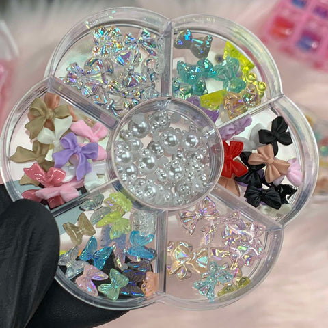 Displayed on a pink, fuzzy background, the black-gloved hand presents the Dynamic Nail Supply's "60 pcs Mix-shapes Resin Charms with Pearls for Nails Art Designer." This floral-shaped organizer gleams with charming resin charms, colorful bows, and iridescent butterfly shapes, making it an ideal source of inspiration for nail art.