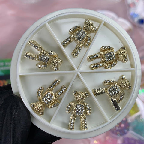 A gloved hand supports a white circular container with six compartments, showcasing the Dynamic Nail Supply's 6 pcs Movable Gold Teddy Bear Charms with Rhinestones for Nails Art Designer, against a soft pink surface. Blurred colorful elements in the background add a touch of whimsy.