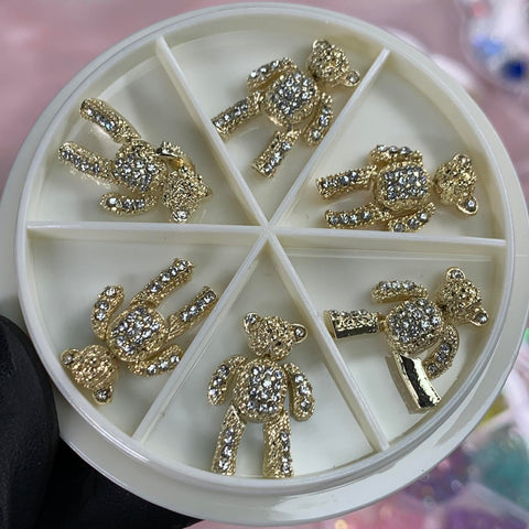Close-up of a round container showcasing the Dynamic Nail Supply product, *6 pcs Movable Gold Teddy Bear Charms with Rhinestones for Nails Art Designer*. Each compartment holds a charm in various playful poses, featuring gold teddy bears adorned with sparkling rhinestones. The slightly blurred background highlights the whimsical appeal of these charming nail art pieces.