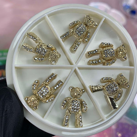 A gloved hand delicately holds a circular white container featuring six sections, each containing a charm from the 6 pcs Movable Gold Teddy Bear Charms with Rhinestones for Nails Art Designer by Dynamic Nail Supply. Set against a blurred pink and green backdrop, these charming bears strike various poses, adding whimsical elegance to any creation.