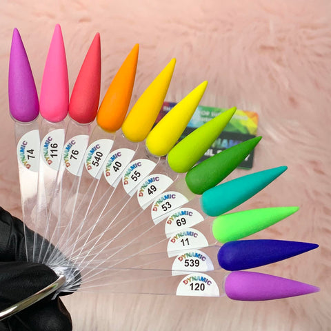 A gloved hand holds the New 2024 Summer Acrylic Collection - Part 01 from Dynamic Nail Supply, featuring ten vibrant, pointed nail color samples on a clear fan. These chip-resistant shades range from pinks to blues and are numbered for easy selection, set against a soft pink background.