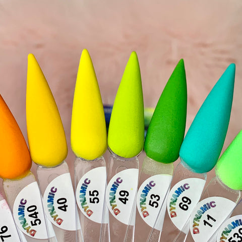 The image features a close-up of seven elongated, pointed nail polish samples arranged in a fan display. Displaying colors from vivid yellow to pink, each sample is marked with a number and the Dynamic Nail Supply brand name. Part of the New 2024 Summer Acrylic Collection - Part 01, these chip-resistant polishes promise durable wear. A softly blurred background highlights their vibrant shades.