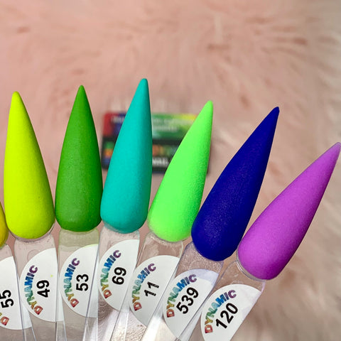 Close-up of seven nail polish color swatches on elongated tips against a fuzzy pink background. The vibrant yellow, lime green, teal, mint green, neon green, deep blue, and vivid purple shades from the "New 2024 Summer Acrylic Collection - Part 01" by Dynamic Nail Supply promise long-lasting nails. Each swatch is numbered and labeled with the brand name Dynamic.