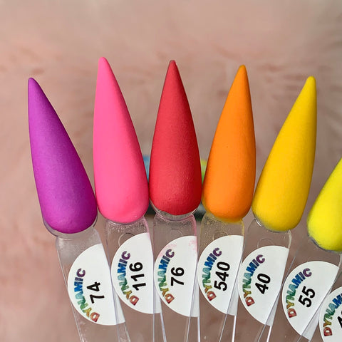 Close-up image of six elongated nail polish display tips from the New 2024 Summer Acrylic Collection - Part 01 by Dynamic Nail Supply, showcasing a range of colors: purple, hot pink, red, orange, orange-yellow, and bright yellow. Each tip is labeled with DYNAMIC and a number: 74, 716, 76, 540, 40, and 55. Perfect for long-lasting nails against a soft pink background.